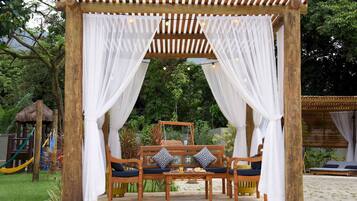 A wooden gazebo with white drapes and a bench with cushions.