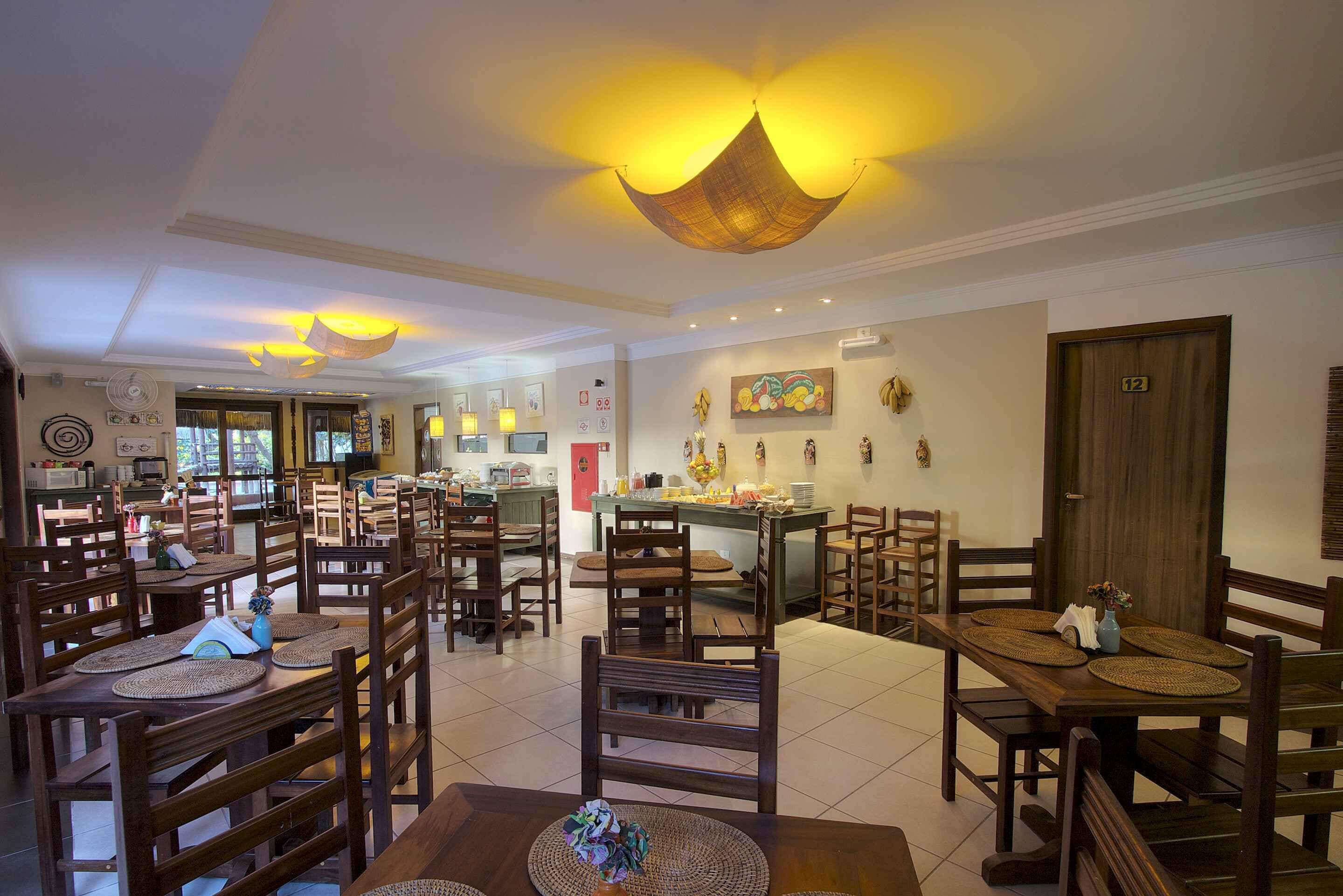A spacious dining area with multiple wooden tables and chairs, a kitchen in the background, and a ceiling with hanging light fixtures.