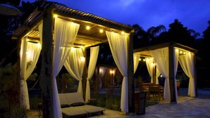 A gazebo with white curtains and string lights, a cushioned seating area, and a stone-tiled patio. - Pousada Porto Mare (São Sebastião)