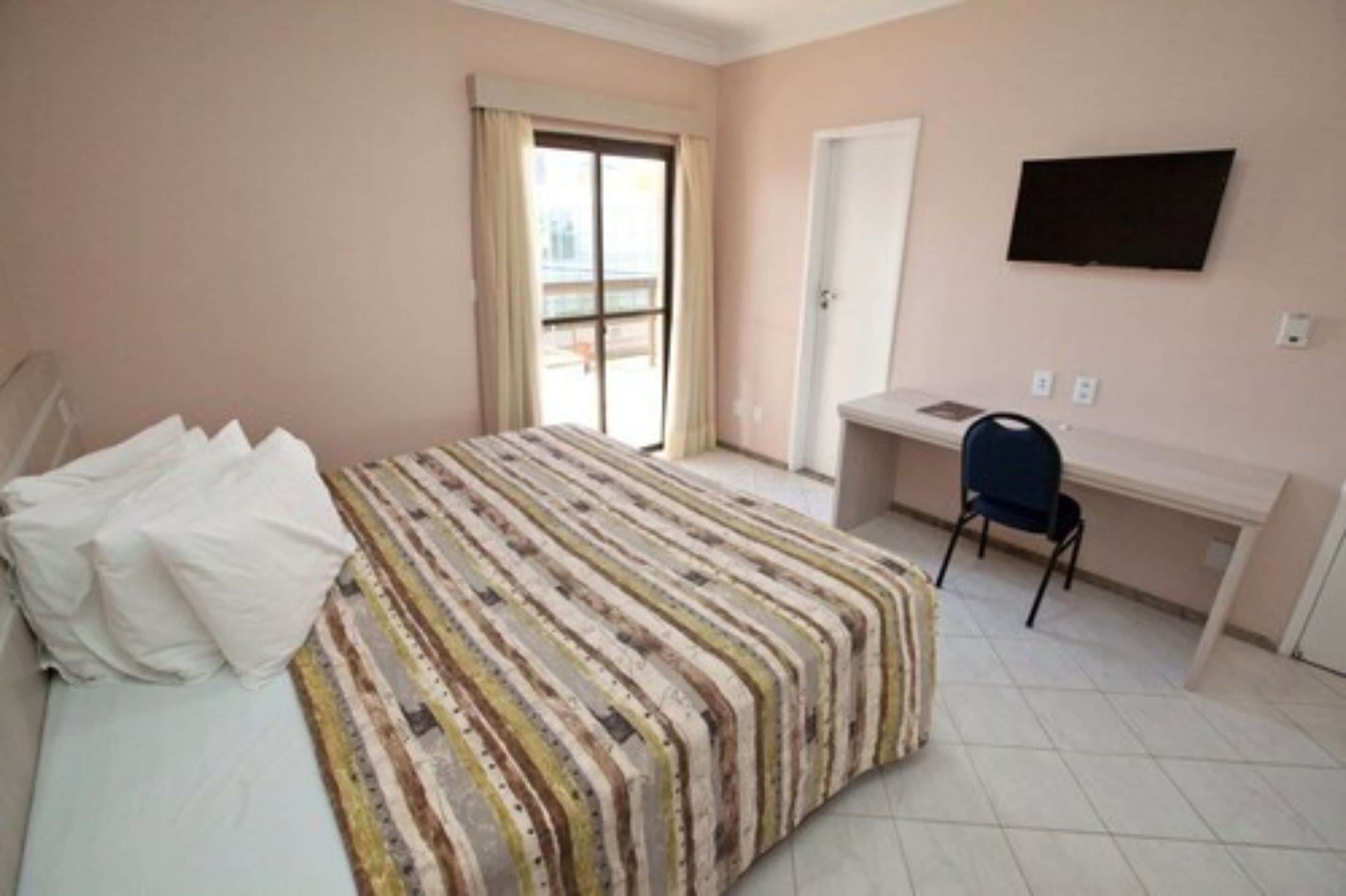 Un dormitorio con cama, escritorio, silla, televisor y puerta corrediza a un balcón.