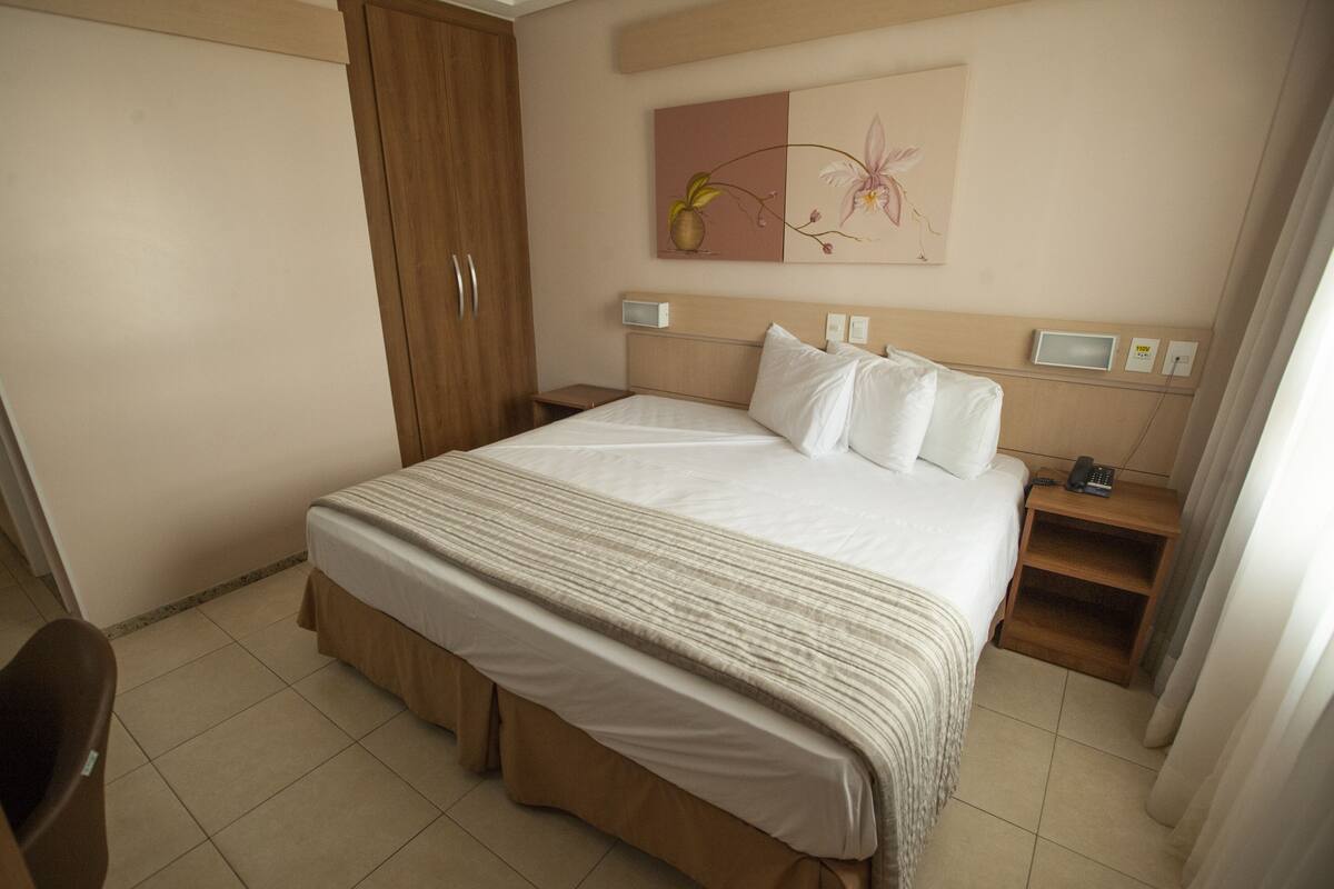 A hotel room with a bed, bedside tables, a nightstand, a wooden wardrobe, and a painting on the wall.
