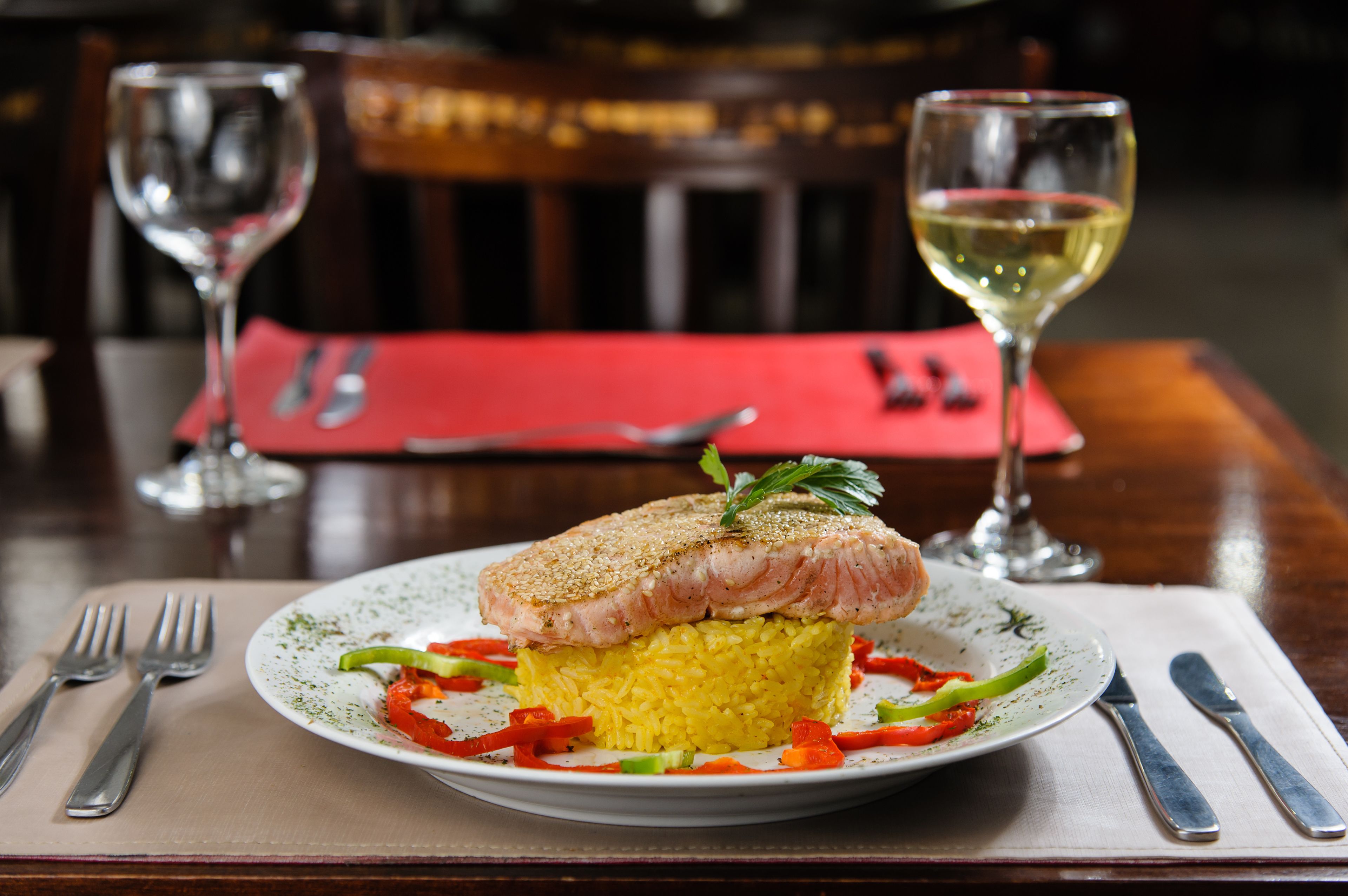 A plate of food with a fish fillet on top of rice, accompanied by vegetables and garnished with herbs. There are wine glasses and cutlery on the table.