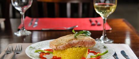 Un plato de comida con un filete de pescado sobre arroz, acompañado de verduras y adornado con hierbas. Hay copas de vino y cubiertos sobre la mesa.