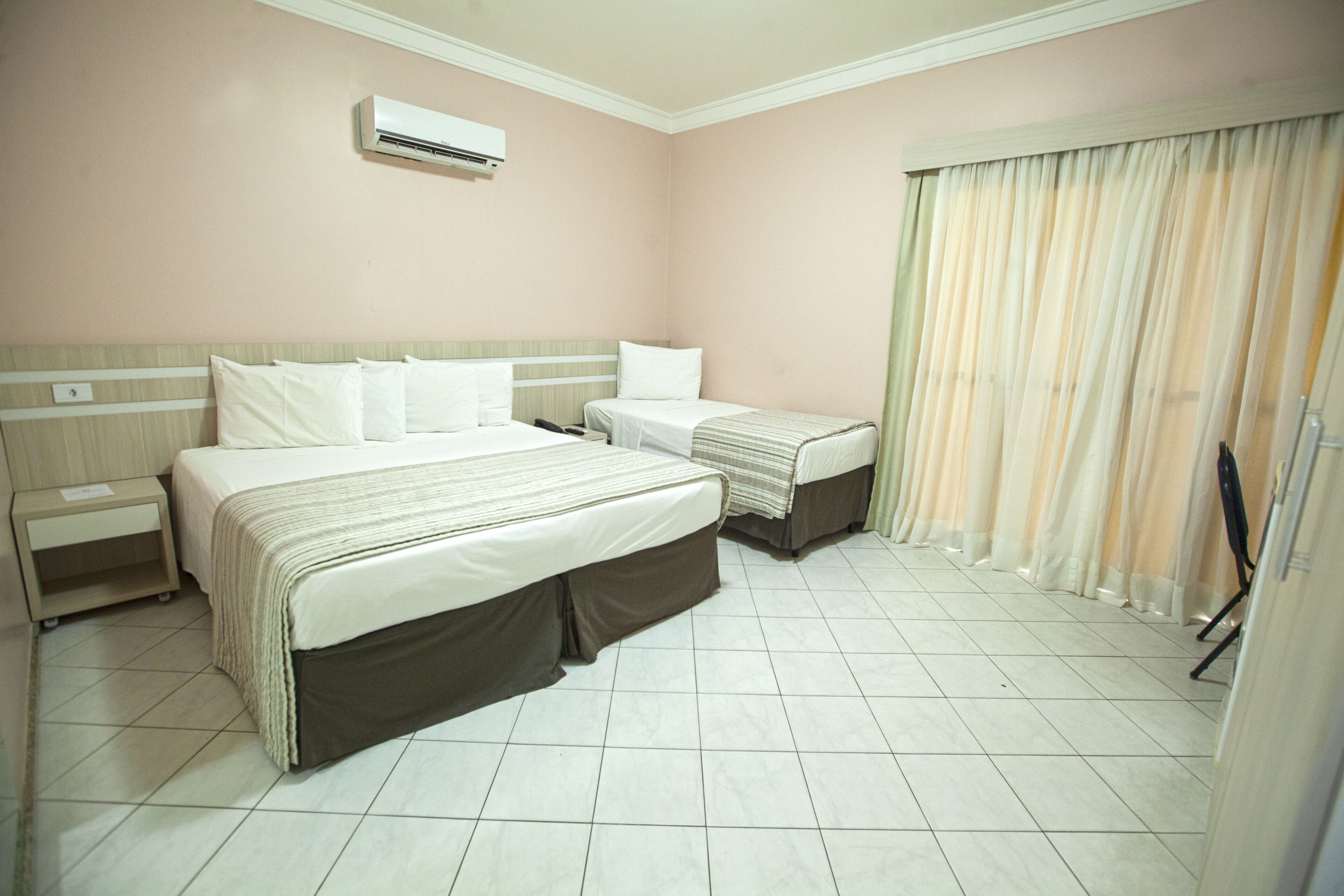 A hotel room with two beds, a nightstand, a window with curtains, and an air conditioning unit.