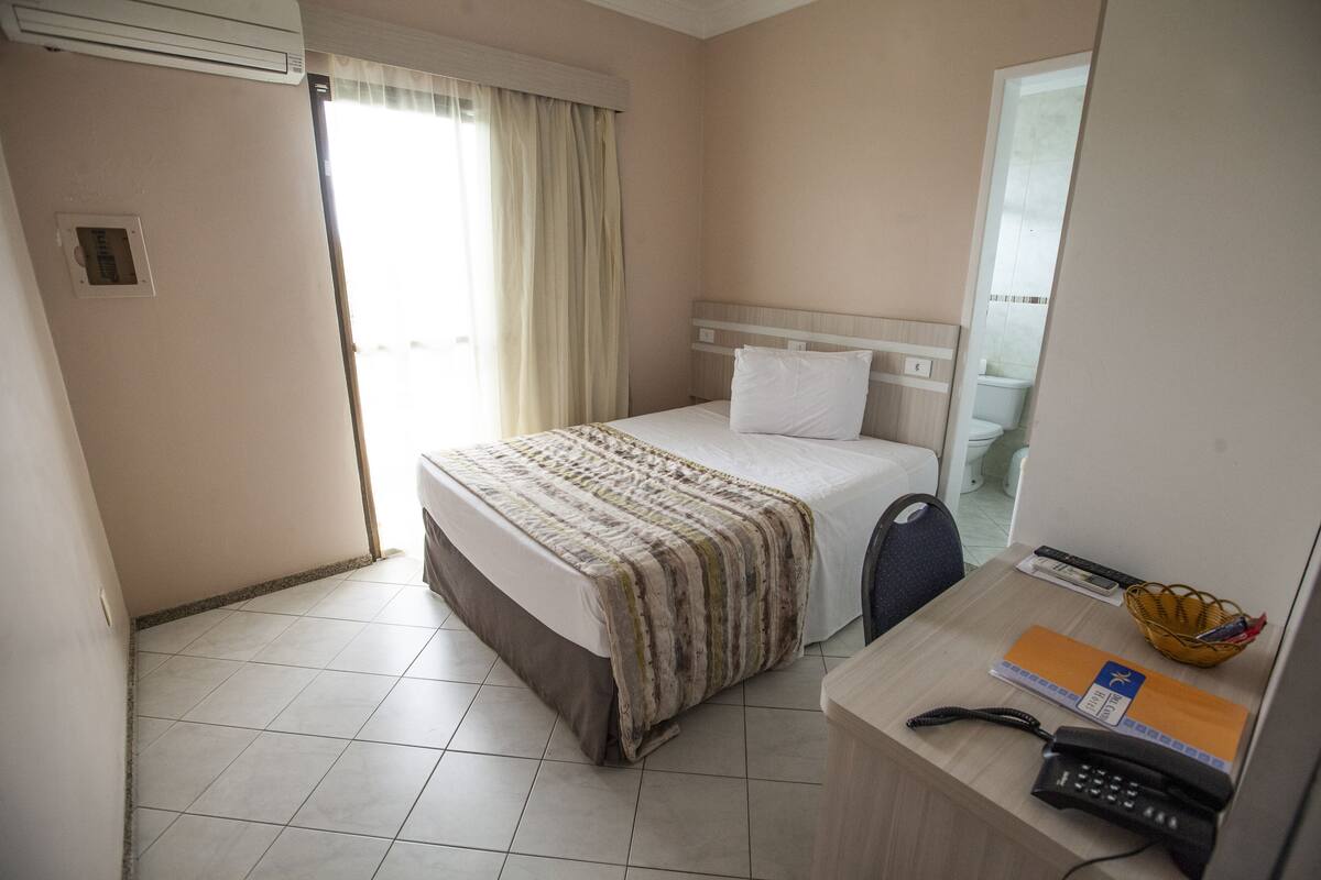 A hotel room with a bed, a desk with a telephone, a basket, and a window with curtains.