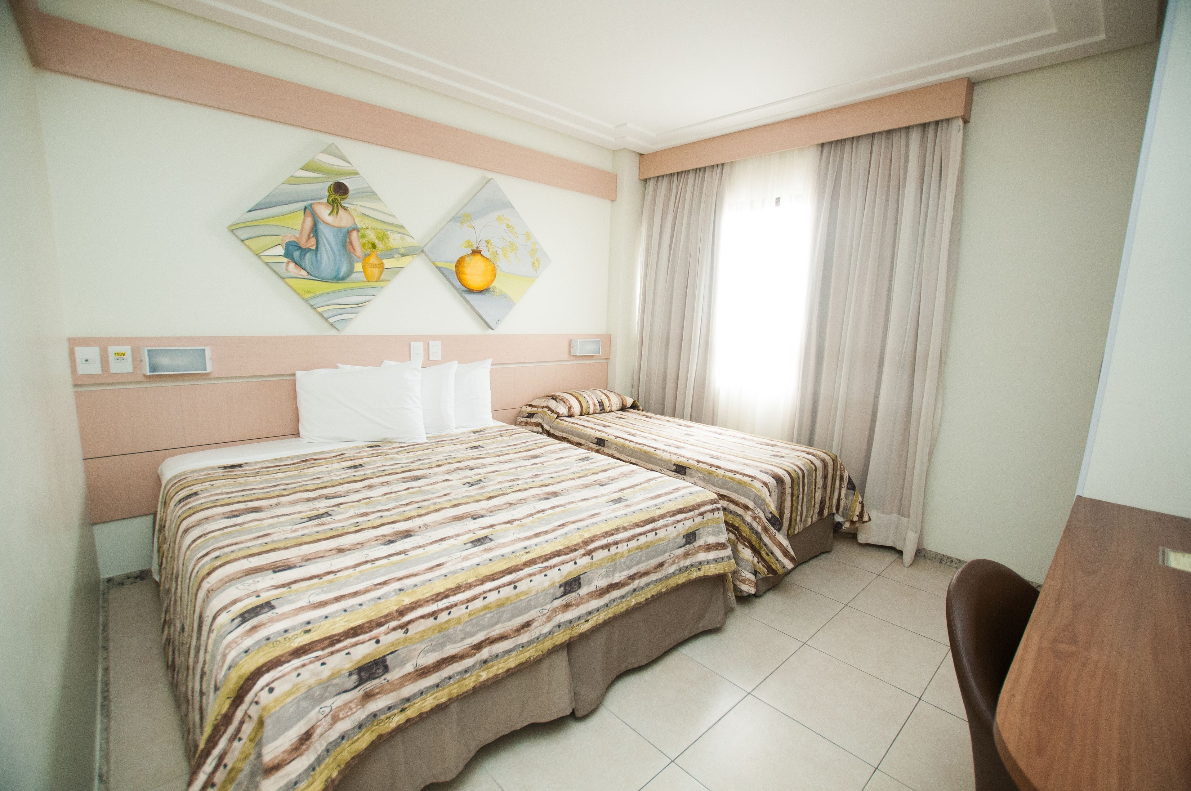 A hotel room with two beds, a desk, and a chair. There are two paintings on the wall, a window with curtains, and a thermostat on the wall.