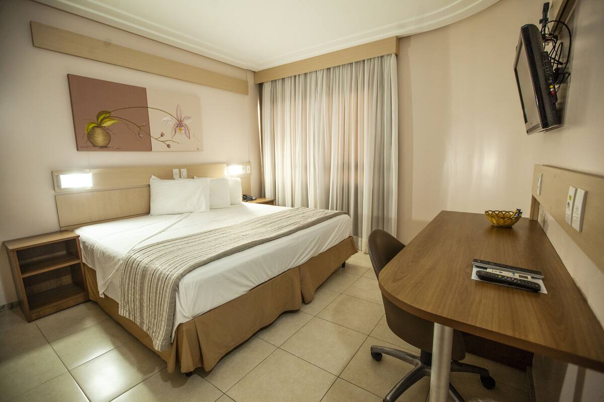 A hotel room with a bed, a desk, a chair, a TV, and a window with curtains.
