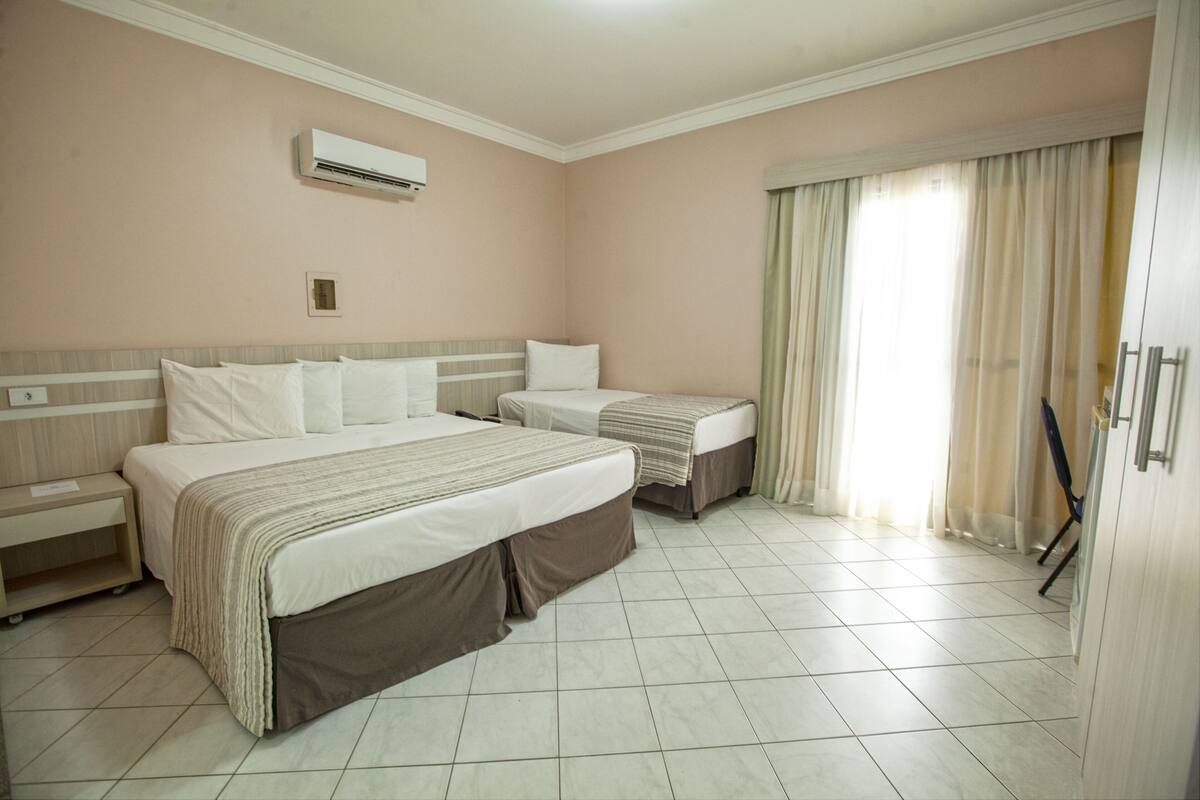 A hotel room with two beds, a nightstand, an air conditioning unit, and a window with curtains.