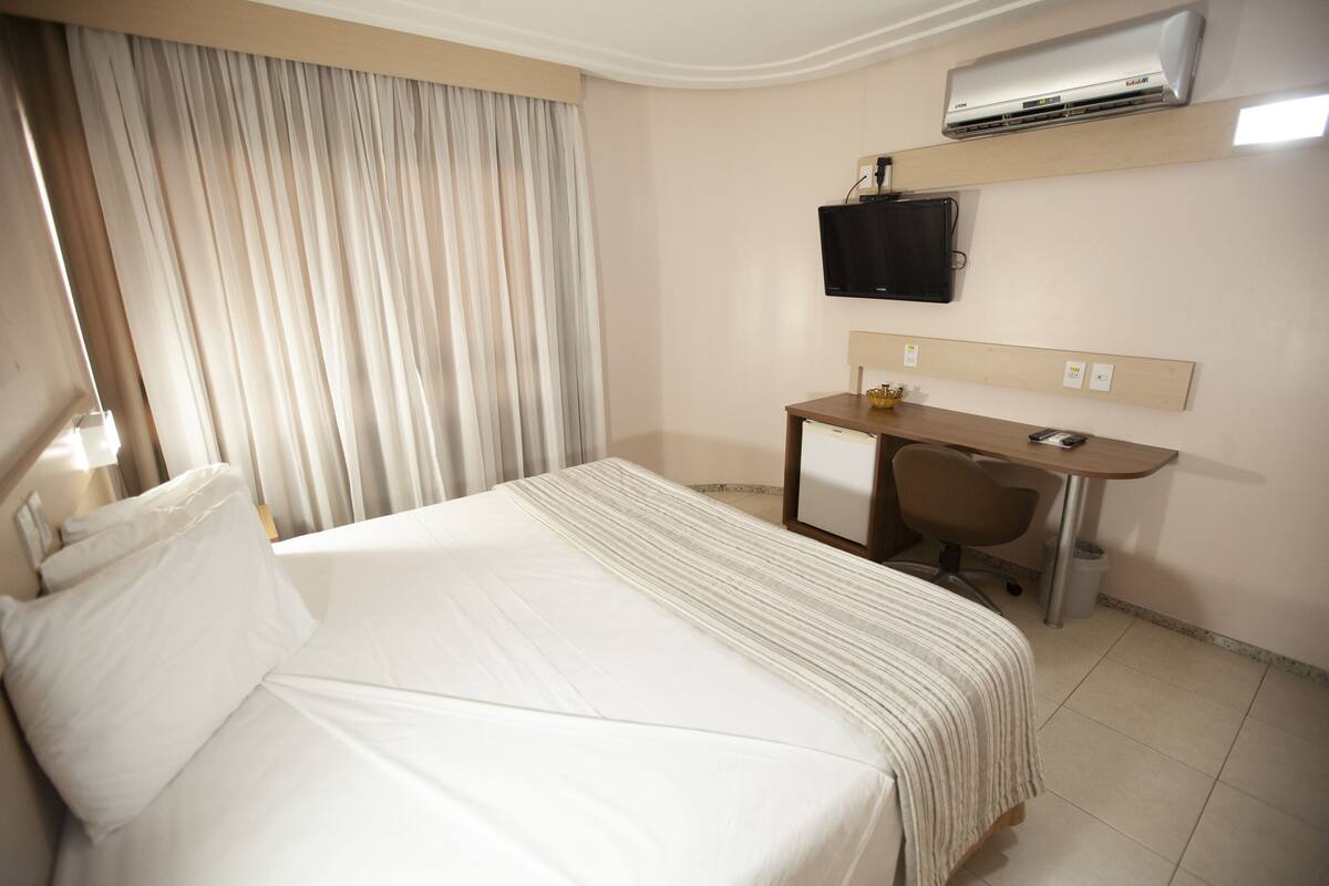 A hotel room with a bed, a desk, a chair, a television, and an air conditioning unit.