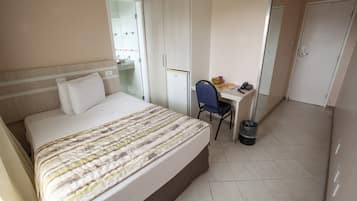 A hotel room with a bed, a desk, a chair, a trash bin, and a wardrobe.