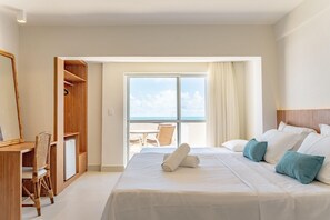 Une chambre d’hôtel équipée d’un lit, d’un bureau, d’une chaise, d’une armoire et offrant une vue sur la mer grâce à une grande fenêtre.