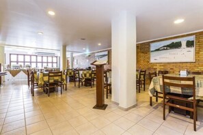 A spacious restaurant with multiple tables and chairs, a brick accent wall, and a large window providing natural light. - Pizzato Praia Hotel (Natal)