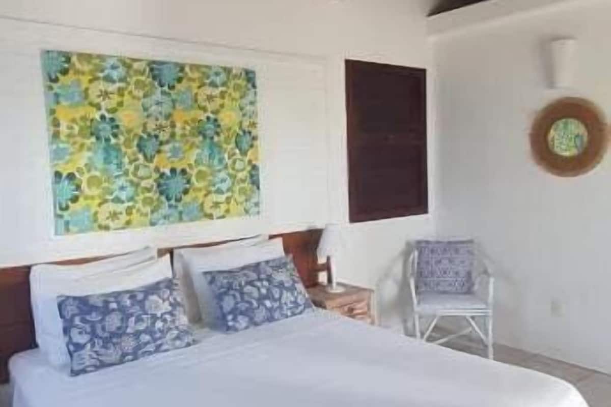 A bedroom with a bed, a hanging light fixture, a wall with a floral painting, and a chair.