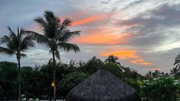 Ein Schwimmbecken mit einer strohgedeckten Struktur, Palmen und einem Sonnenuntergangshimmel.