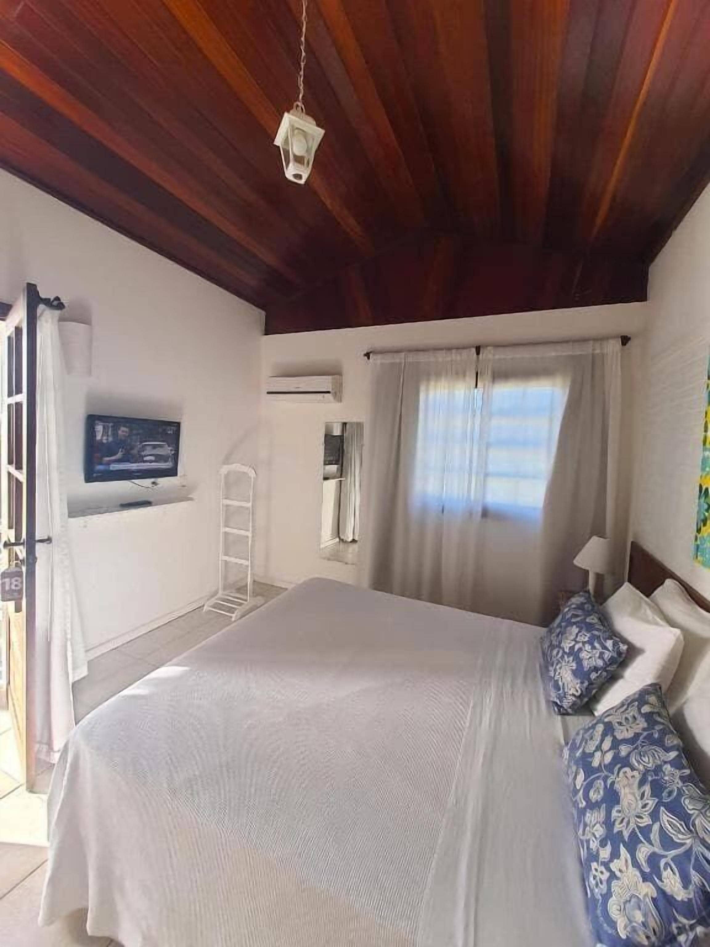 Een slaapkamer met een bed, een aan de muur bevestigde televisie, een raam met gordijnen en een houten plafond.