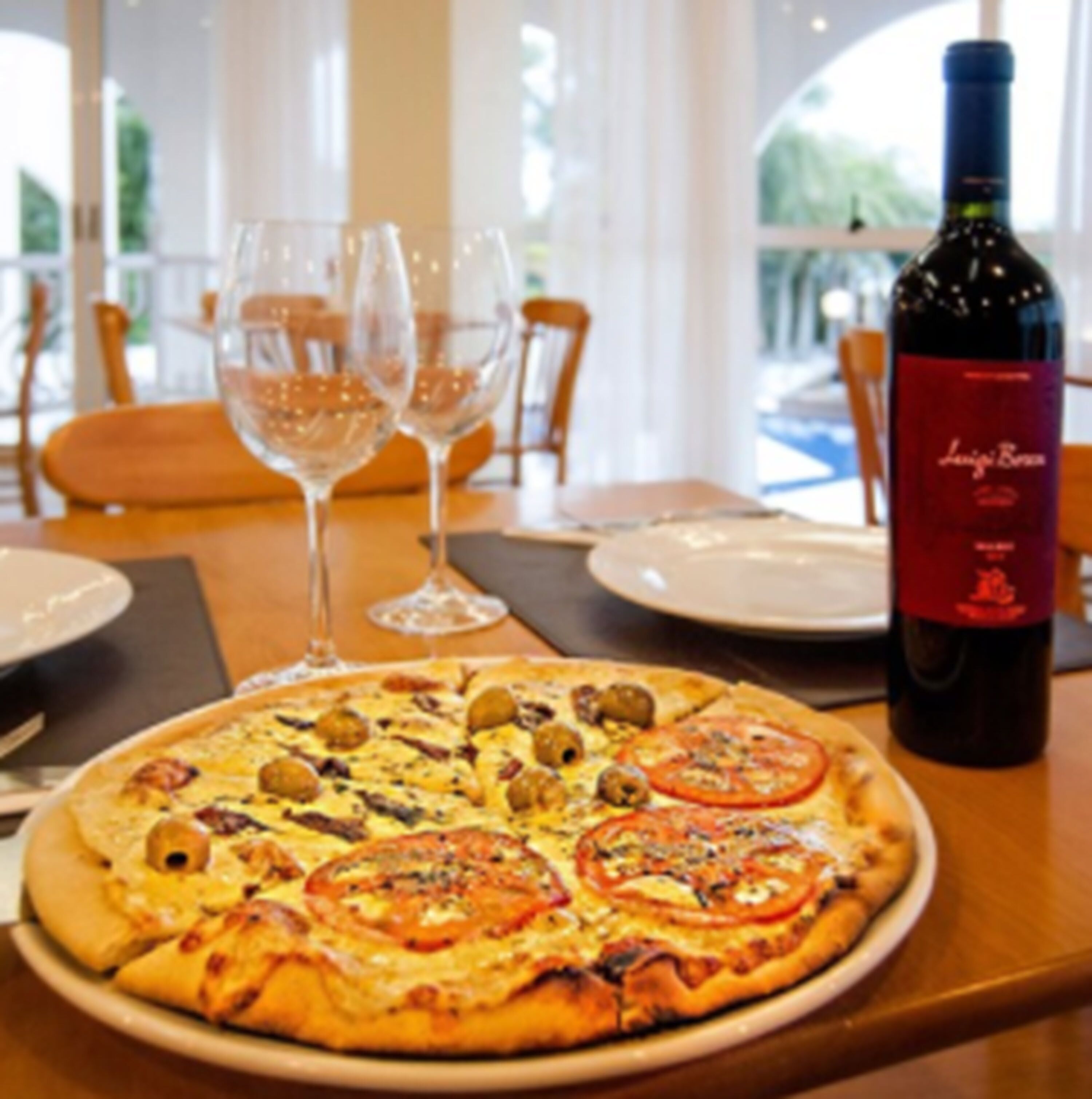 A pizza with toppings on a plate, a bottle of wine, and two glasses on a table.