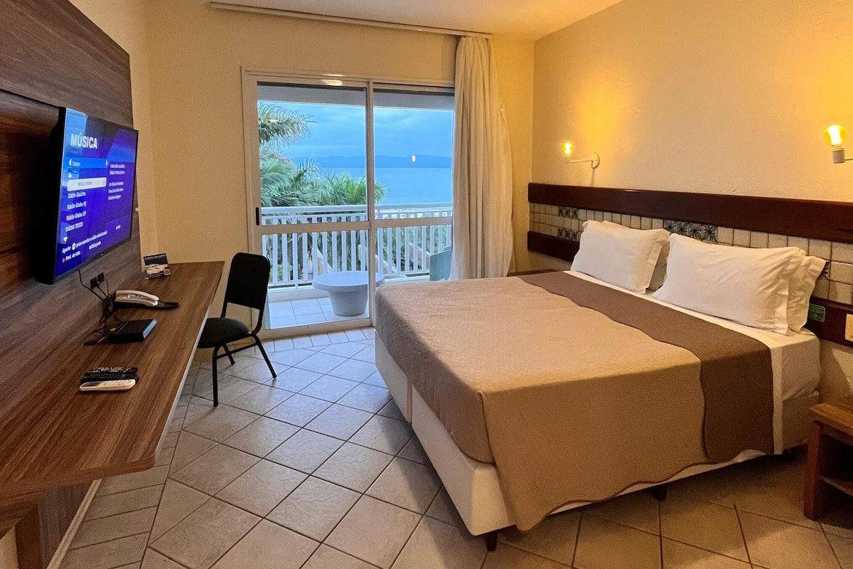 Quarto de hotel com cama, escrivaninha com televisão, cadeira e varanda com vista para o mar.