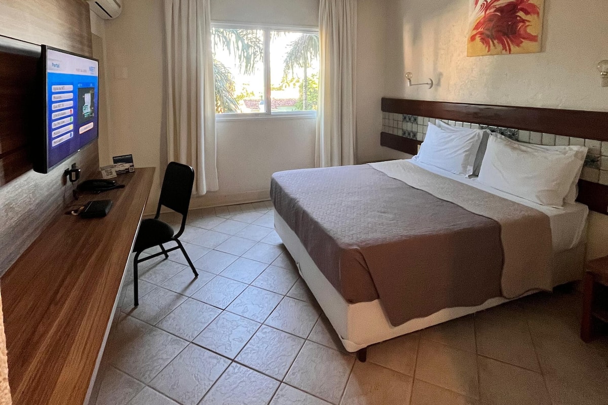 Quarto de hotel com cama, escrivaninha com computador, cadeira e janela com cortinas.