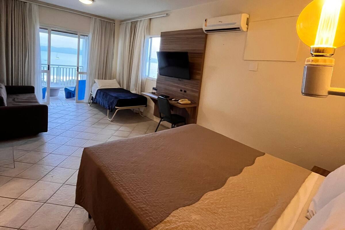 Quarto de hotel com cama, escrivaninha, cadeira, televisão e varanda com cortinas.