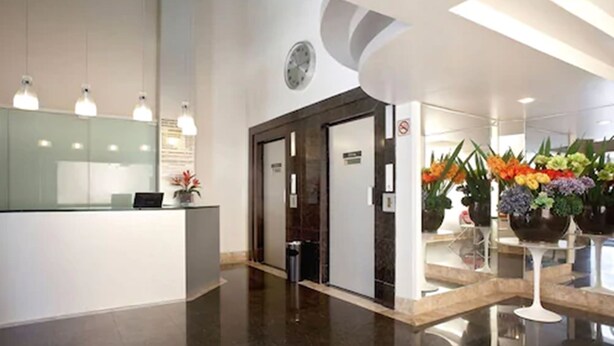 A modern reception area with a glass counter, a clock, and potted plants. - Hotel Boulevard Express (Belo Horizonte)