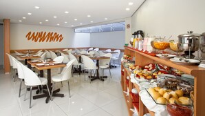 A modern dining area with multiple tables and chairs, a buffet counter with various food items, and a large wall with a unique design. - Hotel Boulevard Express (Belo Horizonte)