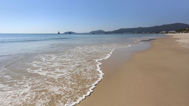 Una playa con aguas cristalinas, una orilla arenosa y montañas a lo lejos.