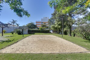 Un terrain de volleyball de sable, entouré de verdure, avec un bâtiment et une enseigne visible au fond.