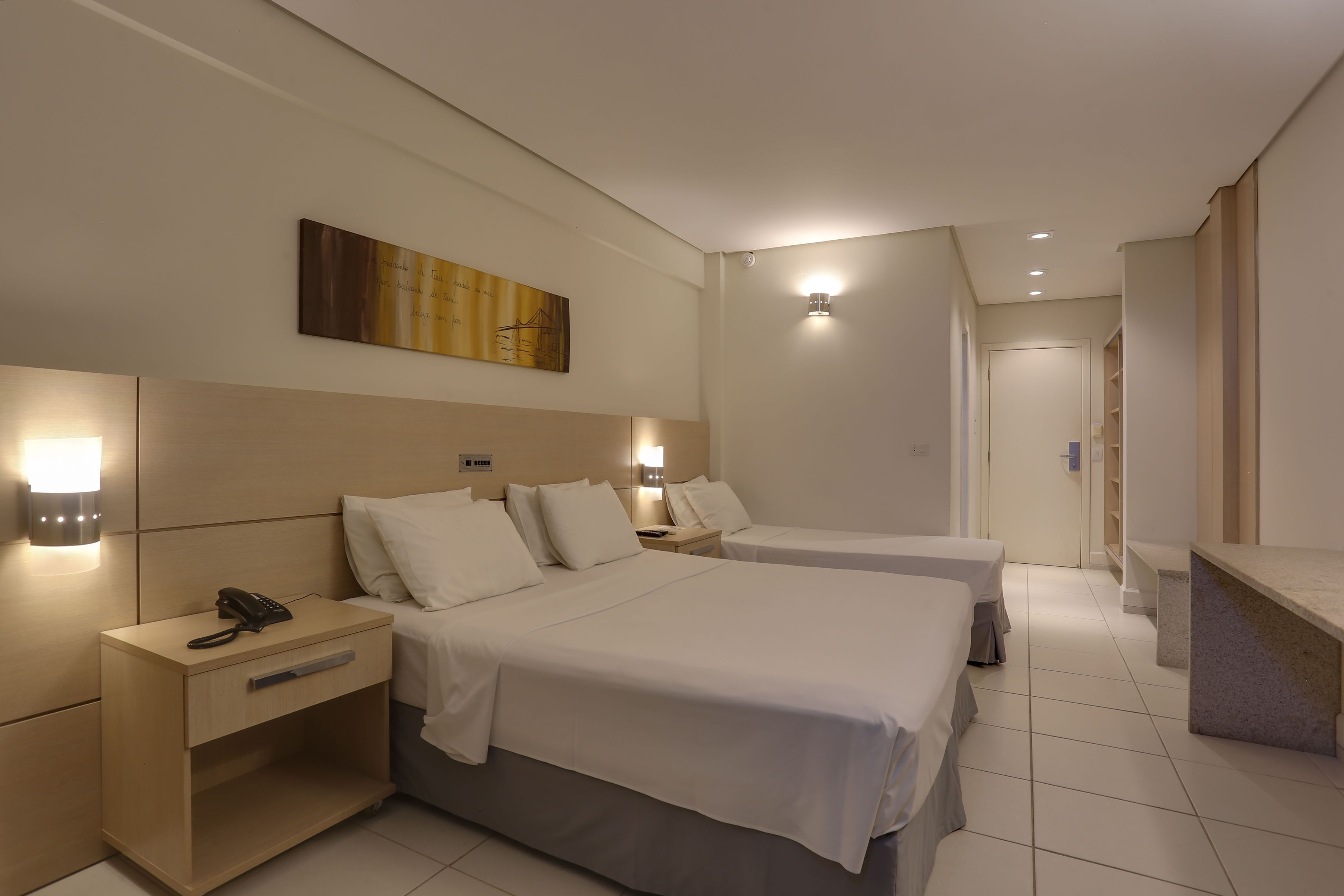 A hotel room with two beds, a bedside table with a phone, a nightstand, and a wall-mounted light fixture.
