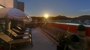 Una terraza en la azotea con una tumbona, una mesa con sombrilla blanca y vistas a la puesta de sol sobre las colinas.