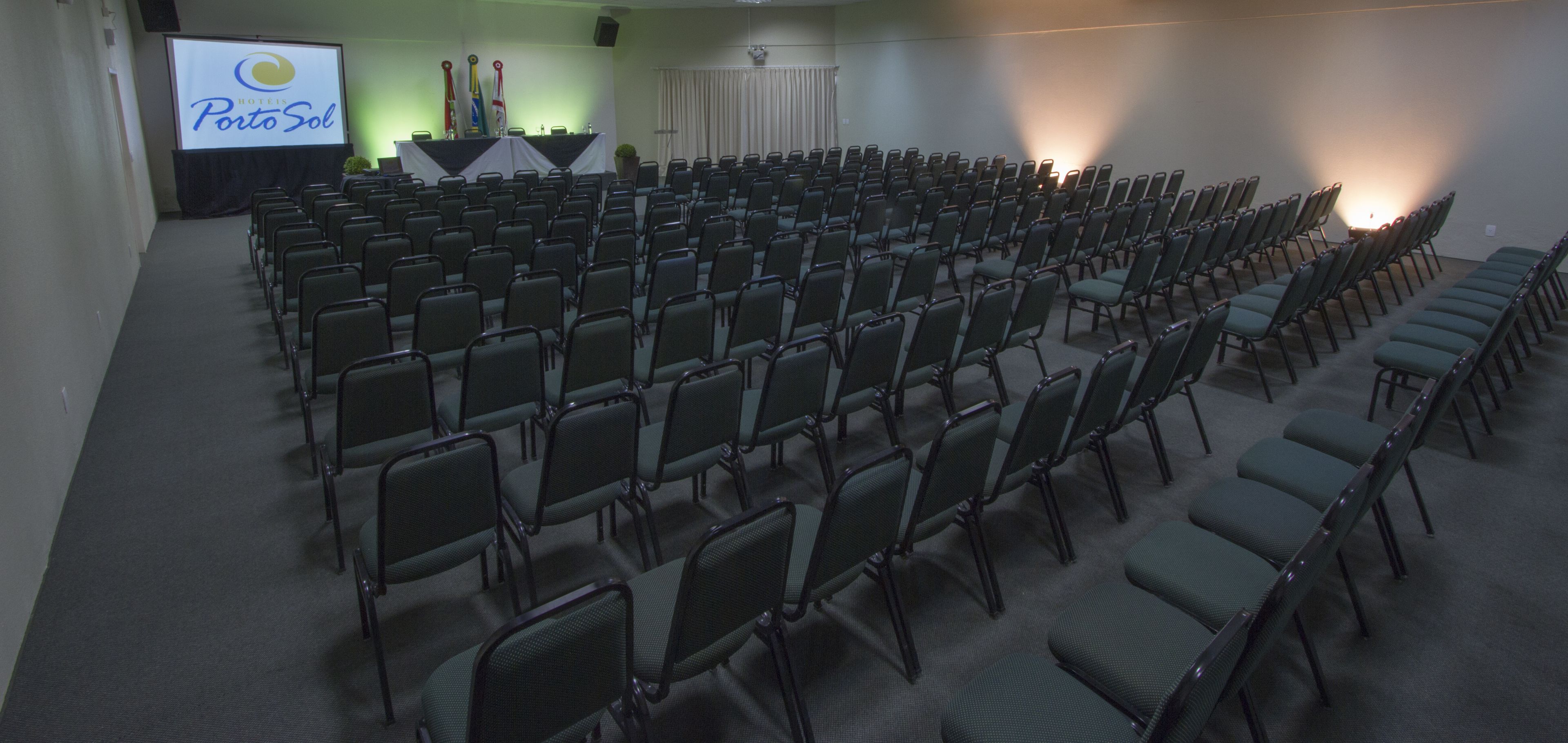 A conference room with rows of chairs, a podium, and a projection screen displaying 'Hotel Porto Sol'.