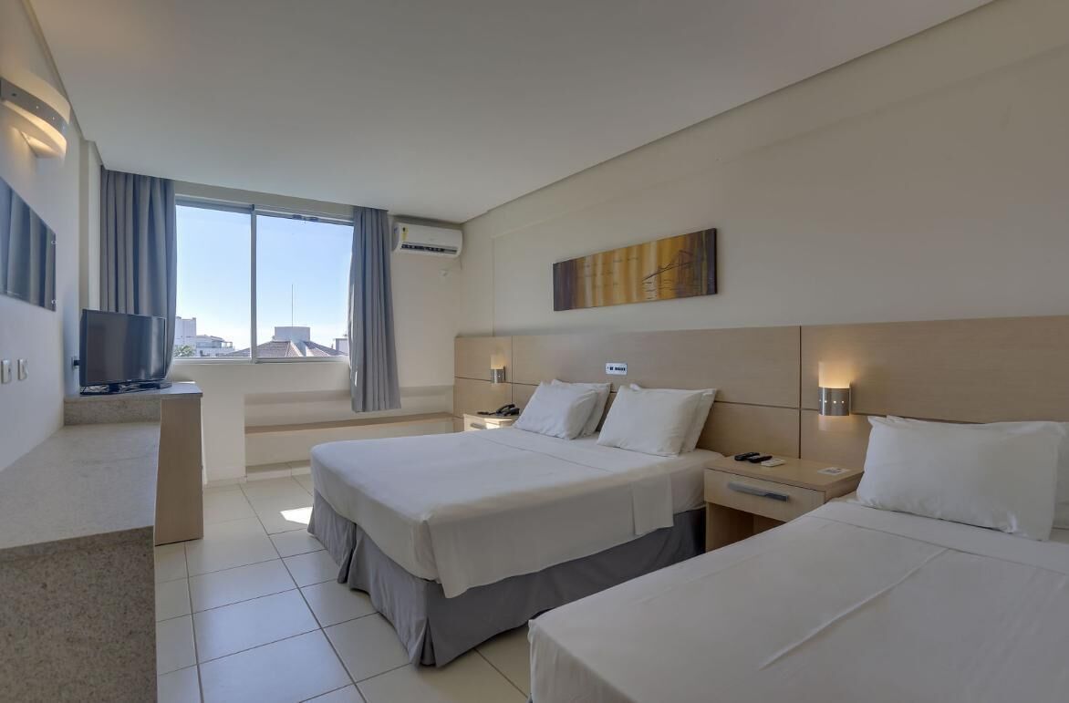 A hotel room with two beds, a TV, a desk, and a large window with a city view.