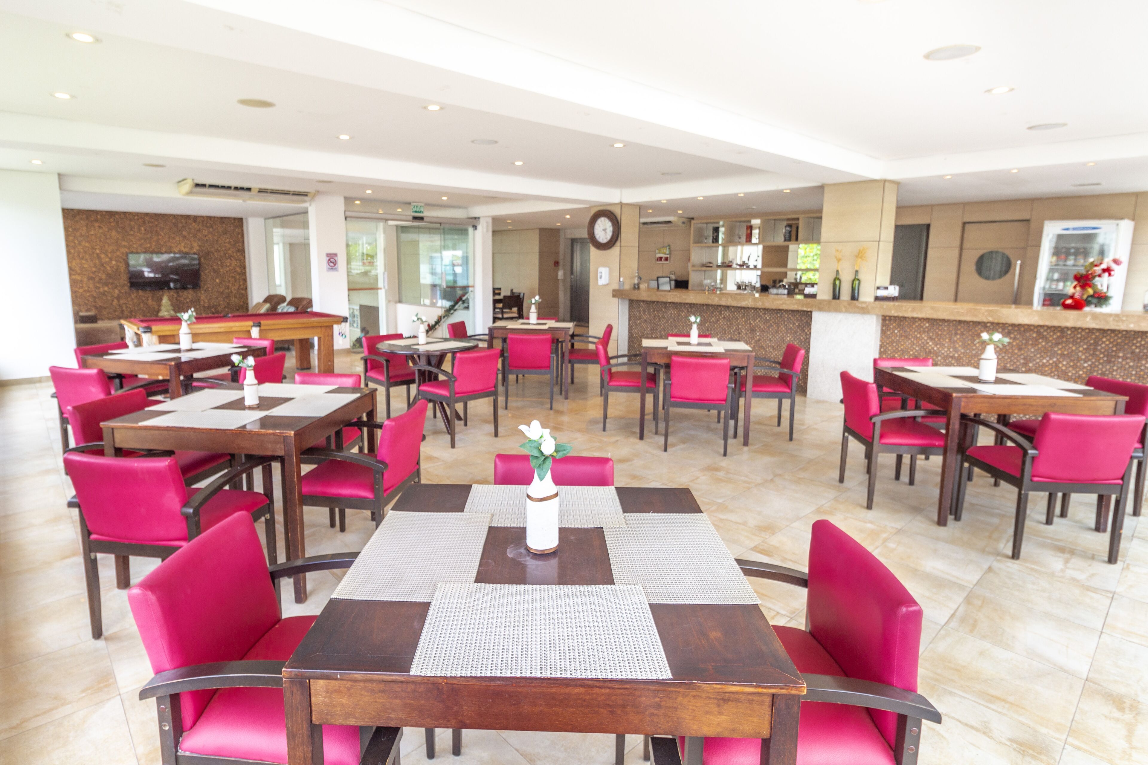A hotel lobby with multiple dining tables and chairs, a reception area, and a clock on the wall.