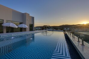 Une piscine extérieure moderne, agrémentée de motifs géométriques en mosaïque, de parasols blancs et offrant une vue sur le coucher de soleil au-dessus d’un paysage vallonné.