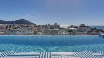 Une piscine à débordement aux motifs géométriques, un ciel dégagé et une vue sur un paysage urbain côtier, avec des bâtiments et une montagne en arrière-plan.