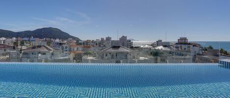 An infinity pool with a geometric pattern, a clear sky, and a view of a coastal cityscape with buildings and a mountain in the background.