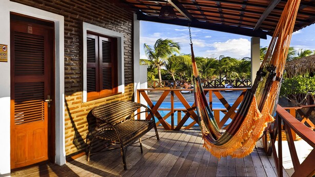 A wooden deck with a hammock, wicker chair, and a view of a pool and palm trees. - Rede Andrade Pousada do Norte (Jijoca de Jericoacoara)