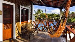 Una terraza de madera con una hamaca, una silla de mimbre y vistas a una piscina y palmeras.