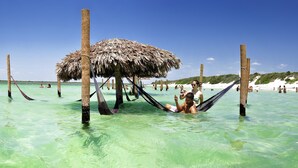 A beach scene with clear turquoise water, a thatched-roof structure, and people relaxing in the water. - Rede Andrade Pousada do Norte (Jijoca de Jericoacoara)