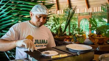 Una persona con gorro y guantes blancos preparando comida en un fogón portátil en un entorno tropical con abundante vegetación.