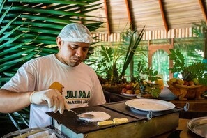 A person wearing a white cap and gloves, preparing food on a portable stove in a tropical setting with lush greenery.