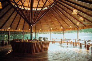 A wooden pavilion with a thatched roof, wooden benches, and a central table with food and drinks.