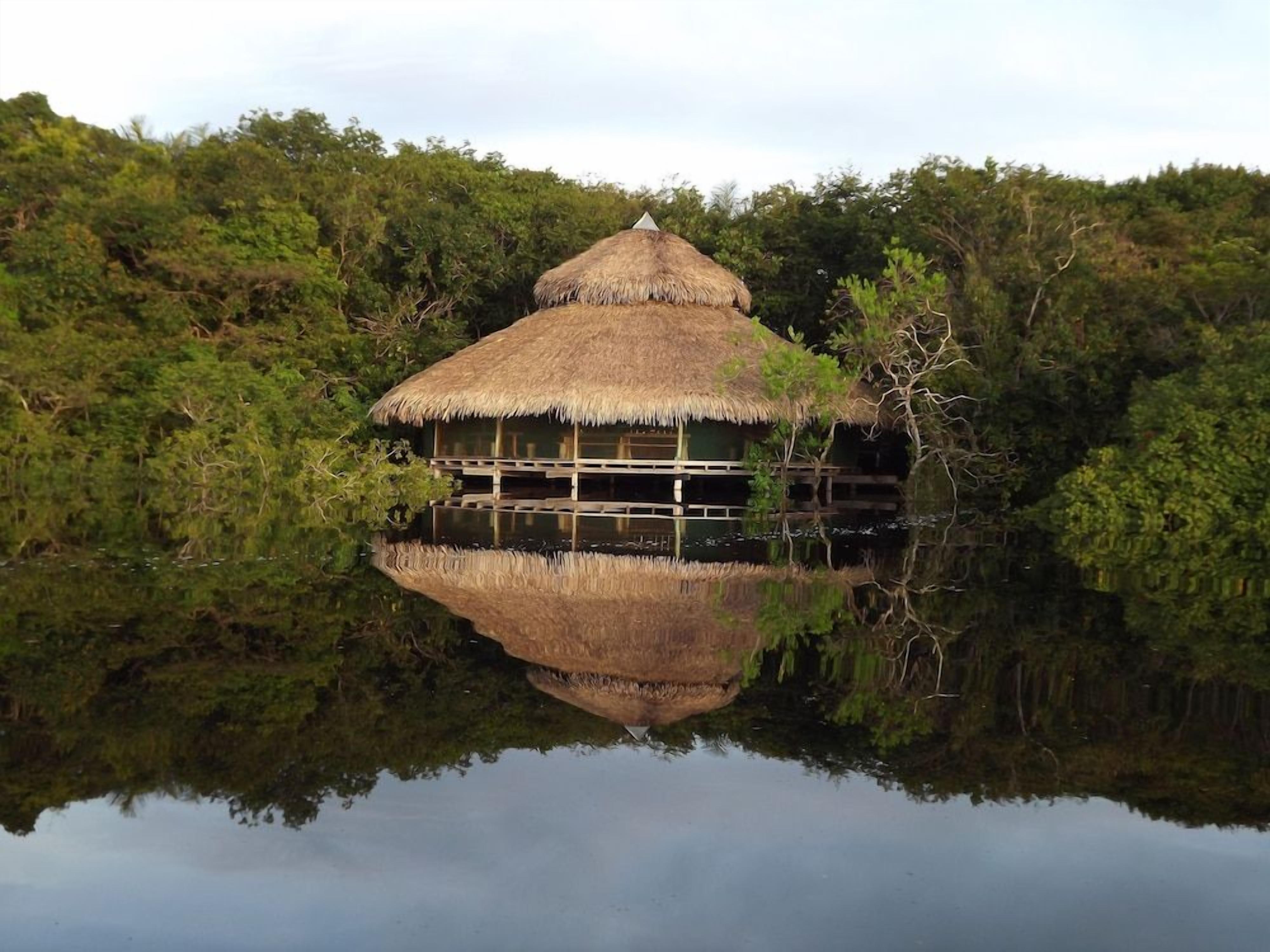 A thatched-roof hut with a reflection in the water, surrounded by dense greenery.