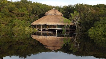 A thatched-roof hut with a reflection in the water, surrounded by dense greenery.
