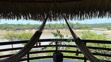 A hammock under a thatched roof overlooking a river and forest.