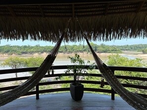 Una hamaca bajo un techo de paja con vistas a un río y bosque.