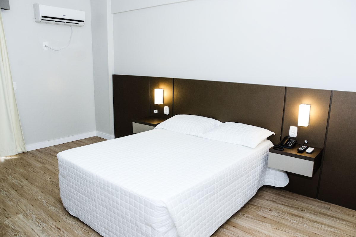 Een net opgemaakt bed met een witte deken, een nachtkastje met een telefoon en afstandsbediening, en een wandairconditioner.