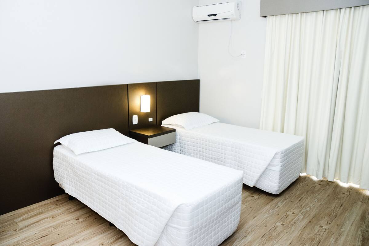 Een hotelkamer met twee eenpersoonsbedden, een nachtkastje, een wandairconditioner en een raam met witte gordijnen.