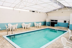 Une piscine intérieure avec un sol carrelé, des chaises blanches et un espace abrité doté d’un toit en métal.