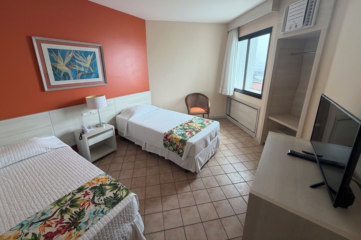 Ein Hotelzimmer mit zwei Betten, einem Sessel, einem Schreibtisch mit Computer, einem Fernseher und einem Fenster mit Vorhängen.