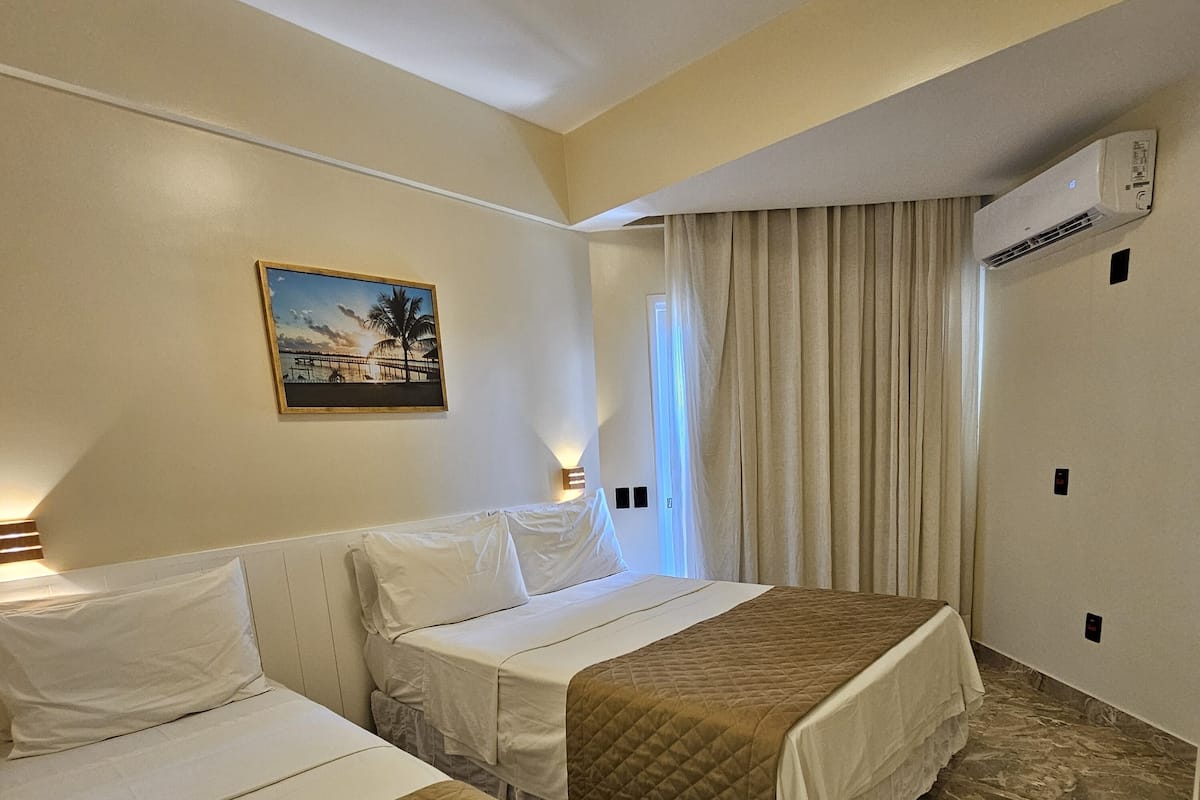 Habitación de hotel con dos camas, un cuadro de una playa y aire acondicionado, sobre un piso de mármol.